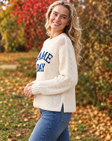 A woman wearing a cream-colored, chunky-knit long-sleeve sweater  with “GAME DAY” embroidered on the front and blue jeans, photographed from behind in an outdoor autumn park with colorful fall foliage.