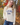 A woman wearing a cream-colored, chunky-knit long-sleeve sweater  with “GAME DAY” embroidered on the front and blue jeans, photographed from behind in an outdoor autumn park with colorful fall foliage.