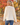 A woman wearing a cream-colored, chunky-knit long-sleeve sweater and blue jeans, photographed from behind in an outdoor autumn park with colorful fall foliage.