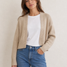 Woman wearing a beige cardigan, white shirt, and blue jeans on a plain background