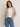 Woman wearing a beige cardigan, white shirt, and blue jeans on a plain background