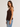 A woman wearing a dark brown ribbed knit tank top and light blue jeans, standing against a plain light gray studio background.
