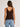 A woman shown from the back wearing a dark brown ribbed knit tank top and light blue jeans against a plain light gray studio background.