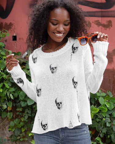 A woman holding sunglasses and a white distressed skull printed knit sweater.