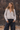 Woman wearing a light oatmeal marled V-neck button-front cardigan with black wide-leg jeans against a rustic wood shingle wall.