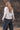 Woman wearing a light oatmeal marled V-neck button-front cardigan with black wide-leg jeans against a rustic wood shingle wall.