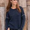 A woman wearing a navy blue knit sweater with silver rope-style trim on the sleeves, standing against a rustic wooden plank wall.