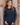 A woman wearing a navy blue knit sweater with silver rope-style trim on the sleeves, standing against a rustic wooden plank wall.