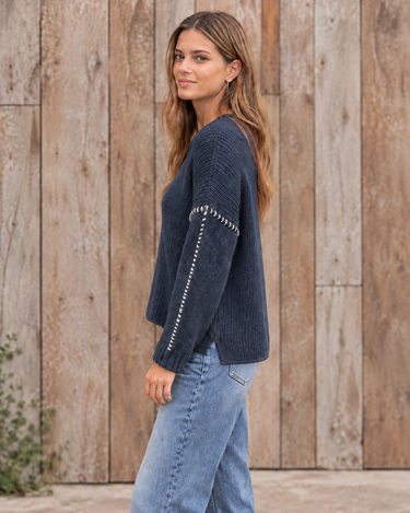 A woman wearing a navy blue ribbed knit sweater with silver contrast stitching on the sleeves, paired with light blue jeans, standing in front of a rustic wooden plank wall.