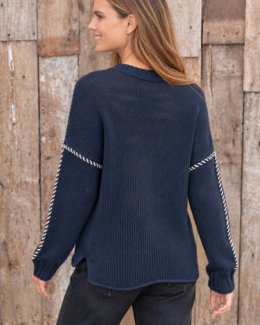 A woman wearing a navy blue knit sweater with silver rope-style trim on the sleeves, shown from the back against a rustic wooden plank wall.