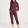 A woman modeling a matching burgundy zip-up jacket and cropped jogger pants set, paired with white sneakers, against a plain light gray studio background.