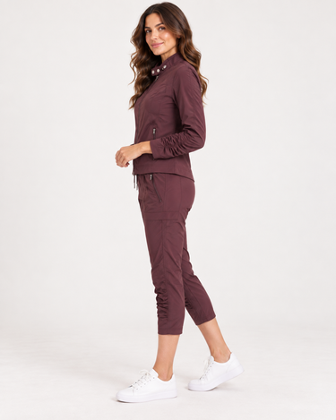 A woman modeling a matching burgundy athletic tracksuit, featuring a zip-up jacket and cropped pants, paired with white sneakers, against a plain white studio background.
