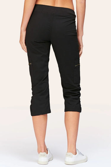 Black cropped cargo pants shown from the back on a model, featuring zippered side cargo pockets and rolled cuffs, paired with white sneakers against a light beige studio background.