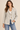 A woman modeling a light beige linen-blend blazer with brown buttons, worn over a white top and blue jeans, against a solid beige background.