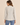 A woman modeling a light beige linen-blend blazer with brown buttons, worn over a white top and blue jeans, against a solid beige background.