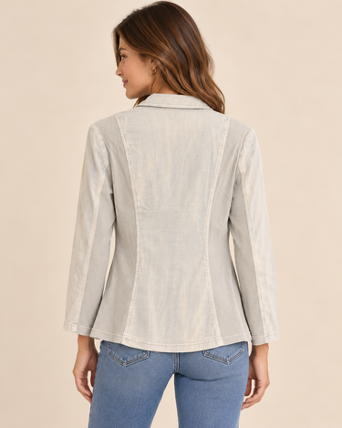A woman modeling a light beige linen-blend blazer with brown buttons, worn over a white top and blue jeans, against a solid beige background.