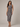 A woman modeling a sleeveless, one-shoulder midi dress with a geometric block pattern in shades of blue, brown, and beige, set against a plain light gray studio background.