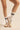 A person's legs wearing white crew socks with black stripes and beige low-top sneakers with gum soles, shown against a warm beige studio background.