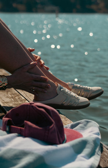 A person sitting by a lake wearing white low-top sneakers, with a burgundy baseball cap resting on a light blue towel in the foreground.