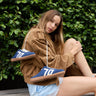 A woman sits on a bench in front of a green hedge, wearing a brown corduroy hoodie and light blue denim shorts, with blue sneakers hanging from her hoodie.