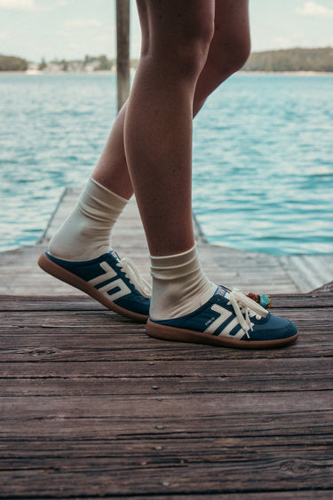 A person standing on a wooden dock by the water wearing blue slip-on shoes with white stripes and cream socks.