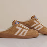 A pair of tan suede slip-on sneakers with white embroidered side stripes and white laces, displayed on a light beige background.