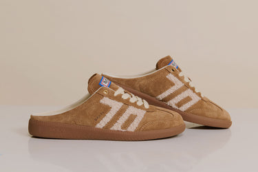 A pair of tan suede slip-on sneakers with white embroidered side stripes and white laces, displayed on a light beige background.