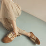 A person wearing beige striped pants and brown suede sneakers with cream lettering, seated against a pale blue floor and cream wall.