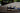 A pair of navy blue and cream New Balance 700 sneakers resting on a weathered wooden deck with a blurred outdoor background.