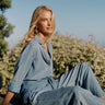 A woman wearing a light blue denim shirt and jeanssits outdoors on a wooden bench with green bushes and a clear blue sky in the background.