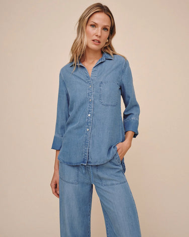 A woman modeling a matching light blue denim two-piece set, featuring a long-sleeve button-up shirt and wide-leg pants, against a solid beige studio background.