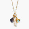 Gold chain necklace with a cluster of charms, including a long baroque pearl, a faceted dark purple iolite gemstone, and a gold floral charm with a small turquoise bead, presented on a white background.