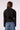 A woman viewed from the back wearing a black cropped jacket with shoulder details and blue jeans, set against a plain light background.