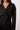 A close-up of a model wearing a black double-breasted cropped trench jacket with dark buttons and shoulder epaulets, paired with a silver necklace against a light gray background.