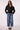 A woman modeling a black belted double-breasted jacket with wide-leg blue jeans and black heels, standing against a light gray studio background.