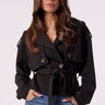 A woman modeling a black belted double-breasted jacket with dark sunglasses and blue jeans, standing against a plain light background.