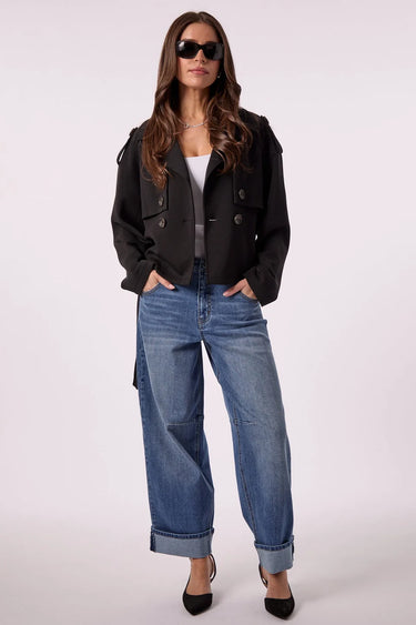 A woman modeling a black cropped double-breasted jacket, blue wide-leg jeans, a white top, and black heels against a light gray studio background.