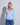 A blonde woman wearing a light blue v-neck sweater and blue jeans, standing against a light gray wall with a soft shadow cast to the side.