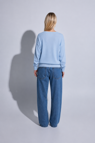 A woman stands with her back to the camera wearing a light blue long-sleeve top and wide-leg blue jeans against a light gray studio background with a prominent shadow.