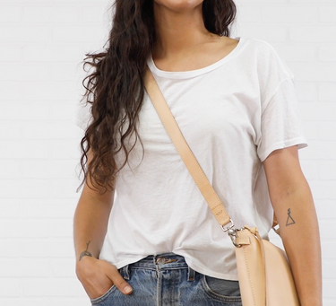 A woman wearing a white crewneck t-shirt and blue jeans, carrying a light beige leather crossbody bag with a silver-toned clasp over her shoulder, set against a white background.