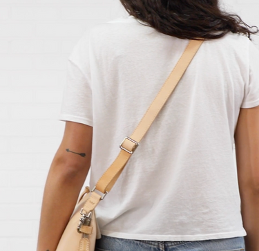 A person wearing a white t-shirt and blue jeans, shown from the back, carrying a light beige leather shoulder bag with a matching strap and silver hardware.