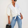 A woman modeling a cream-colored, oversized button-down shirt tied at the waist, paired with blue denim jeans, set against a plain white studio background.