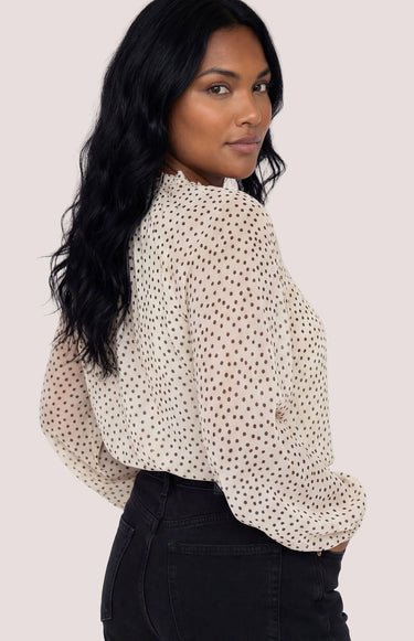 A woman wearing a black polka dot and cream blouse with black jeans on a white background.