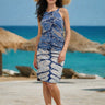 Woman in a blue tie-dye dress and straw hat standing on a beach with turquoise water and straw umbrellas in the background.