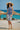 Woman in a blue tie-dye dress and straw hat standing on a beach with turquoise water and straw umbrellas in the background.