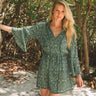 woman wearing floral dress holding onto a tree standing on a sandy beach

