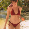 Woman wearing a merlot  halter bikini with an O-ring detail and tie-side bottoms, standing on a sandy beach next to clear water and lush green trees, holding an apple.
