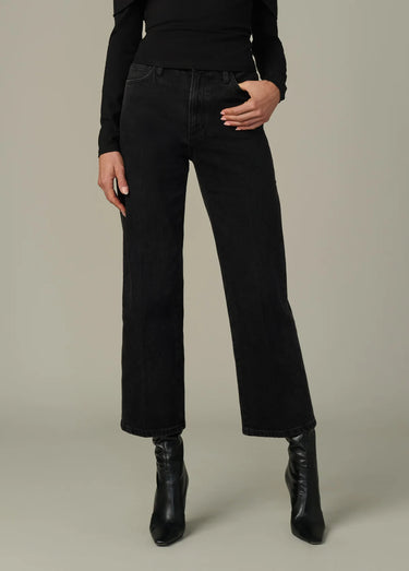 Full-body view of a person wearing black high-rise wide-leg cropped jeans, a black long-sleeve top, and black heeled ankle boots, posing on a light gray background.