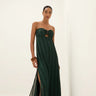 Full-length image of a woman wearing a dark green strapless maxi dress with subtle vertical stripes, a central bust cutout with a knotted detail, and high side slits. She is posing confidently against a white background, wearing brown sandals and wooden bracelets.