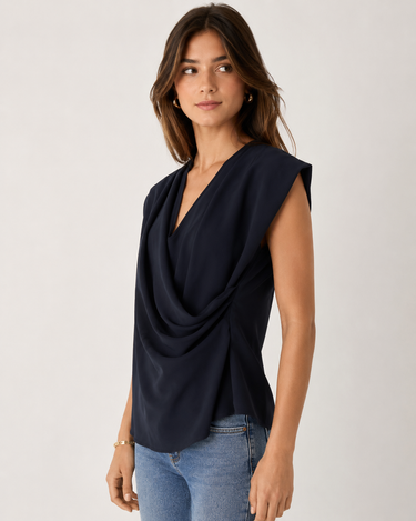 A woman wearing a sleeveless navy blue draped wrap-style top and light wash blue jeans, standing against a plain light gray studio background.